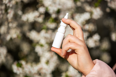 hand holding white bottle of nasal spray in front of blurry white flowers