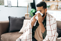 man wrapped in blanket on couch blowing nose